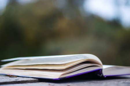 Open book with pages flipping in the wind. Reading on the lake wooden dock on autumn day. Learning enjoying tranquility of nature. Peaceful outdoor environment. Selective focus. Blurred background.の写真素材