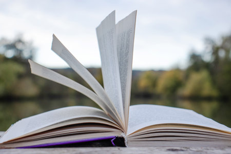 Open book with pages flipping in the wind. Reading on the lake wooden dock on autumn day. Learning enjoying tranquility of nature. Peaceful outdoor environment. Selective focus. Blurred background.の写真素材