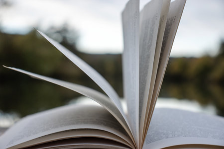 Open book with pages flipping in the wind. Reading on the lake wooden dock on autumn day. Learning enjoying tranquility of nature. Peaceful outdoor environment. Selective focus. Blurred background.の写真素材