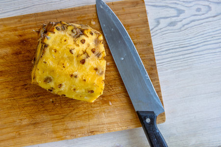 A partially peeled pineapple sits on a wooden cutting board next to a large kitchen knife. Fresh,...