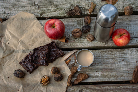 Piece of handmade brownie cake on the baking paper, metal thermos and mug with hot milk coffee, tee or cacao, red apple, nuts. Picnic on the wooden pier or terrace. Lunch break. Flat lay. Top viewの写真素材