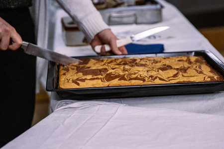 Woman cutting a piece of a tasty marble pie with a big knife on black baking tray. Selling cakes at the festival. Party food on the big table covered with white tablecloth. Selective focus.の写真素材