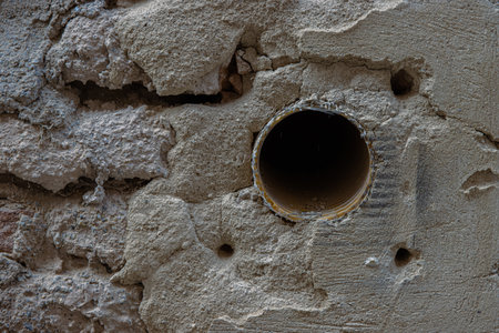 Old damaged shabby red brick wall with hole. Brick texture of an old building with scratches and cracks. Metal tube sticking out of cracked stucco. Grunge background. Close up. Copy spaceの写真素材