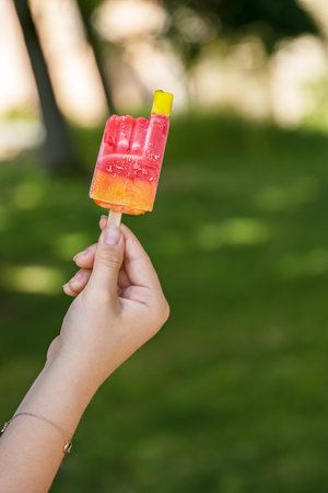 Close-up of a vibrant colorful red yellow orange ice cream with popsicle stick held in a hand of unrecognizable child. Sunny summer day in park. Blurred background. Copyspace. Selective focusの写真素材