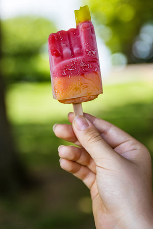 Close-up of a vibrant colorful red yellow orange ice cream with popsicle stick held in a hand of unrecognizable child. Sunny summer day in park. Blurred background. Copyspace. Selective focusの写真素材