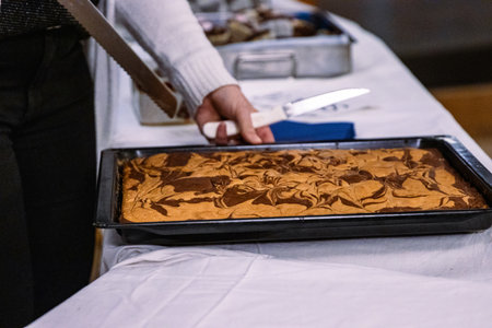 Woman cutting a piece of a tasty marble pie with a big knife on black baking tray. Selling cakes at the festival. Party food on the big table covered with white tablecloth. Selective focus.の写真素材
