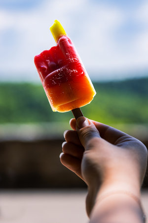 Hand holding red fruit popsicle ice cream on the wooden stick. Strawberry, cherry, orange, mango flavor. Sunny summer day in park. Blurred background. Copy space. Selective focusの写真素材