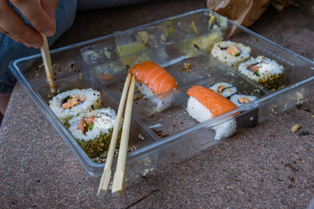 Young woman wearing jeans is sitting on the stone steps eating sushi rolls out of opened half-empty plastic tray. Hands holding chopsticks. Eaten Sushi set, food sticks, wasabi and ginger packets.の写真素材