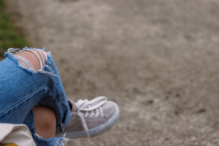 Young girl wearing ripped blue jeans with big hole on the knee is sitting on the bench crossed her legs. Woman in destroyed distressed denim. Close up of textile texture. Selective focus, blurredの写真素材