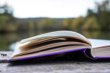 Open book with pages flipping in the wind. Reading on the lake wooden dock on autumn day. Learning enjoying tranquility of nature. Peaceful outdoor environment. Selective focus. Blurred background.の写真素材