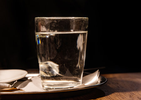 Close up of glass with hot water served on the metal tray with white napkin, sugar pack and tea bag against black background. Transparent tea cup on the table. Copyspaceの写真素材