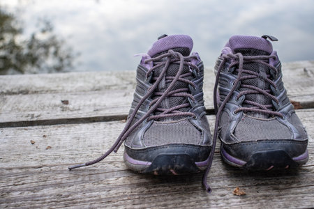 Closeup of a pair of gray and purple hiking shoes on a wooden dock by water. Old worn out torn vintage sport boots on a deck of the lake beach. Active lifestyle outdoors near on a peaceful autumn day. Selective focus.の写真素材