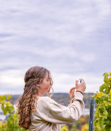 A young woman with long curly hair capturing scenic vineyard landscape views with her smartphone. Teenager blogger girl photographing autumn nature with her phone. Copyspaceの写真素材