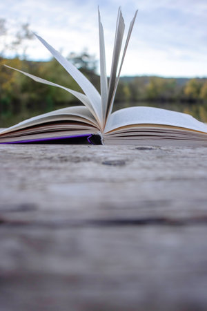 Open book with pages flipping in the wind. Reading on the lake wooden dock on autumn day. Learning enjoying tranquility of nature. Peaceful outdoor environment. Selective focus. Blurred background.の写真素材