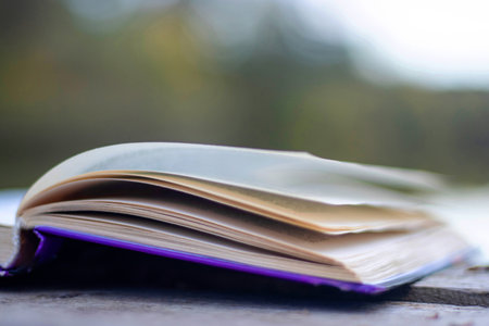 Open book with pages flipping in the wind. Reading on the lake wooden dock on autumn day. Learning enjoying tranquility of nature. Peaceful outdoor environment. Selective focus. Blurred background.の写真素材