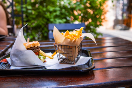 Fast food meal at a cozy outdoor cafe. Classic burger with crispy fries served on the tray. Cheeseburger with beef patty, fresh lettuce and sesame seed bun and golden French fries in a metal basket.の写真素材