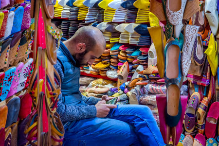 FEZ, MOROCCO - DECEMBER 10: Man playing with phone and selling colorful leather slippers in Fez old town. December 2016のeditorial素材