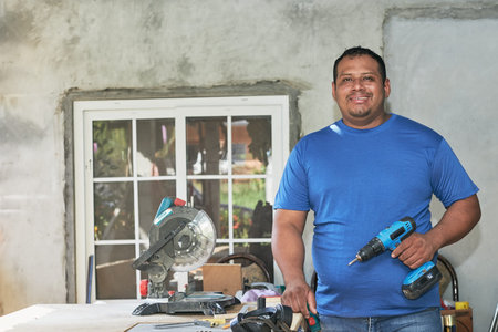latin man smiling, holding cordless drill in cabinetry workshop