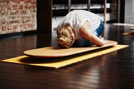 Middle aged woman resting on child pose yoga board in yoga class. Female yogi doing balasana...