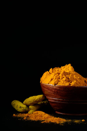 Turmeric powder and roots or sticks in wooden bowl on black background