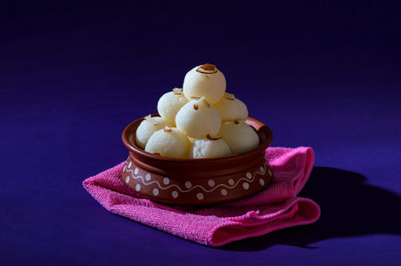 Indian Sweet or Dessert - Rasgulla, Famous Bengali sweet in clay bowl with blue napkin on violet...