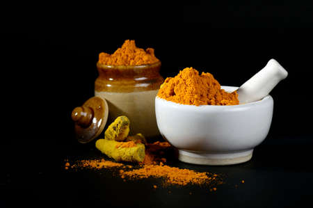 Turmeric powder in mortar with pestle and roots with clay pot on black backgroundの写真素材