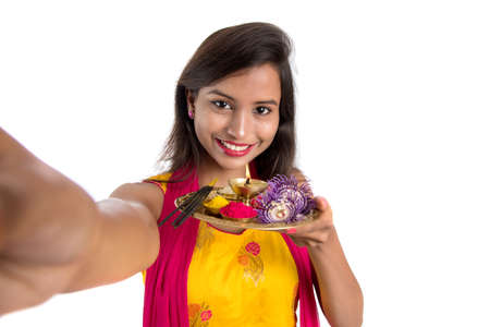 Beautiful young happy girl taking selfie with pooja thali using a mobile phone or smartphone on a white backgroundの写真素材