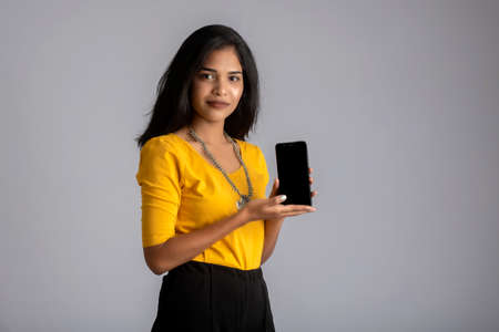 Young beautiful girl holding and showing blank screen of smartphone or mobile or tablet phone on a gray background.の写真素材