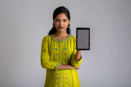 Young beautiful girl holding and showing blank screen of smartphone or mobile or tablet phone on a gray background.の写真素材