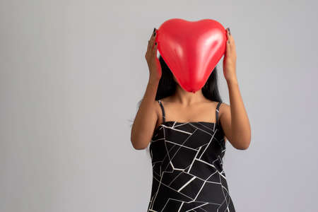 Cheerful cute girl in fancy outfit posing with heart shape balloon on grey backgroundの写真素材