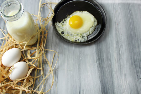 Overhead view of white eggs in egg cups surrounded by straw,  milk and a fried egg in an iron...