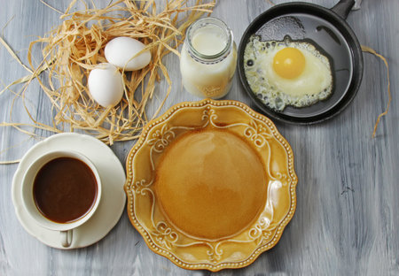 Overhead view of a table set with a plate, eggs, milk in a bottle, black coffee and fried egg in a pan on a grey textured backgroundの写真素材
