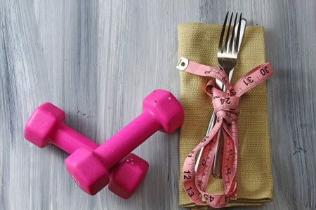 A napkin with fork and knife wrapped up in a measuring tape with pink dumbbells on a grey textured...