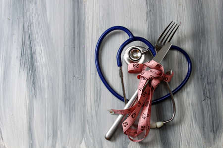 Concept of healthy eating with a fork and a knife wrapped in a measuring tape and a blue stethoscope in the form of a heart on a grey wooden textured background with copy space.の写真素材