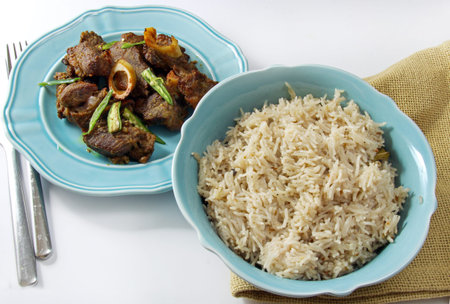 Mutton fry on a plate with a bowl of aromatic basmati rice on grey background.