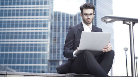 Front view of a handsome young businessman wearing glasses and a suit sitting on the stairs and working with his laptop. Downtown.の写真素材