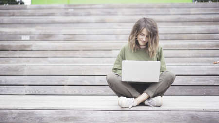 Woman sitting on the bench while typing on laptop. Wooden background. Front view.の写真素材