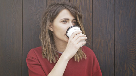 Portrait of young woman drinking coffee to go while standing near a wooden wall.の写真素材