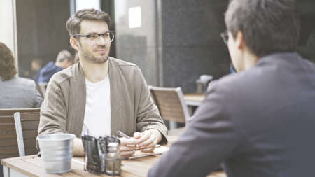 Two friends are smiling talking having a conversation at table of the cafe outdoors. A man dressed in a jacket wearing eyeglasses listening to his friend talkingの写真素材