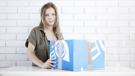A young disappointed woman opening a blue cardboard box with a box cutter at home delivered gift by...