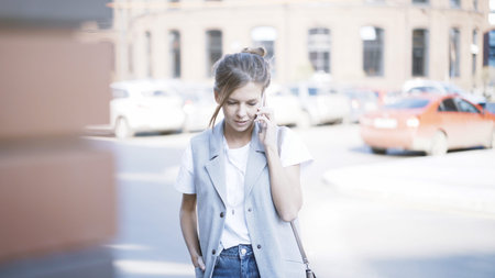 A young smiling pretty girl dressed in a jacket is having a call outside outdoors