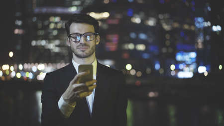 A face of successful businessman dressed in a black suit wearing eyeglasses is texting a message on his phone using an app at night timeの写真素材