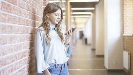 Young girl wearing a white shirt and jeans is looking at her smartphone screen standing in a business building lobby.の写真素材