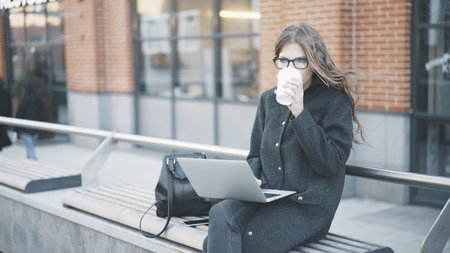 Calm young businesswoman in glasses and a coat drinking coffee and holding her laptop sitting...