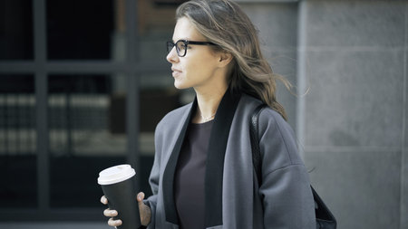 Woman wearing glasses and a gray coat is holding a cup of coffee and smilng. A downtown background....