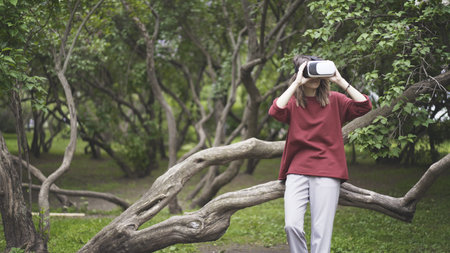 A young girl dressed in a red sweatshirt and gray pants having fun in in virtual reality glasses sitting on a tree trunk in the parkの写真素材