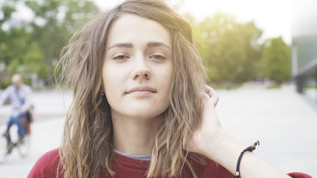 A young cute casual brunette girl holding hair in a red sweatshirt outside in the morning trees on the backgroundの写真素材