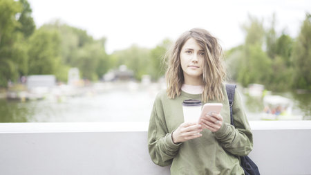 A serious young girl is listening to a music outdoors dressed in a green sweatshirt drinking a cup of coffeeの写真素材