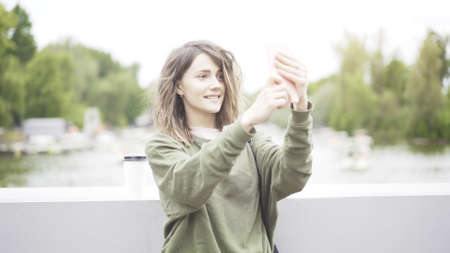 A young smiling woman is taking a selfie outdoors dressed in a green sweatshirt drinking a cup of coffee in the park near the waterの写真素材