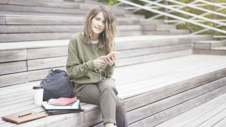 A young pretty smiling girl dressed in a green sweat shirt is listening to music outdoors sitting on the stairs texting looking at the cameraの写真素材